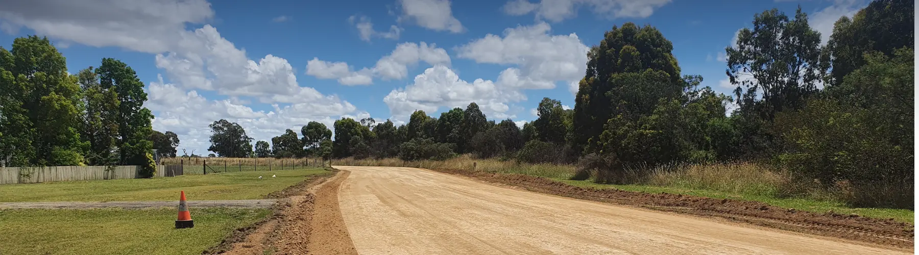 Newly formed gravel rural road curving past paddocks and gum trees, with a traffic cone at the edge